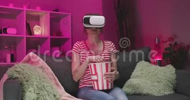 年轻女子戴V R耳机，晚上用爆米花<strong>看电影</strong>。 坐在沙发上的女人戴着VR眼镜